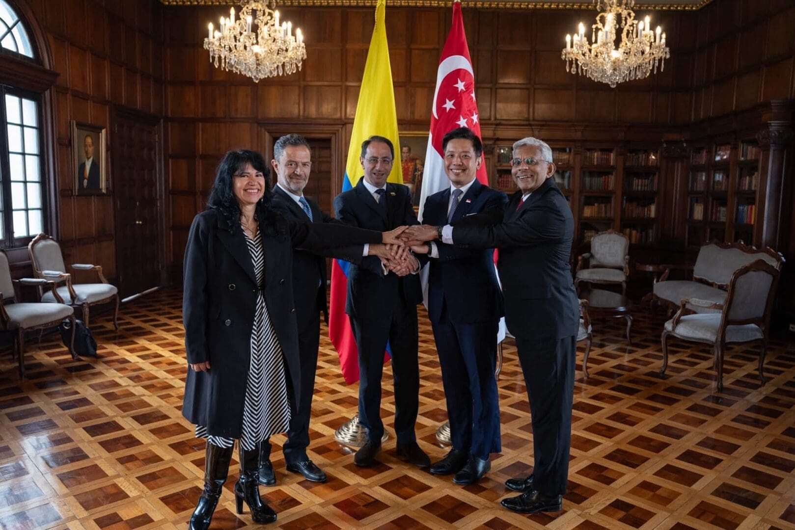 Five officials pose together with their hands joined in front of the Colombian and Singaporean flags.