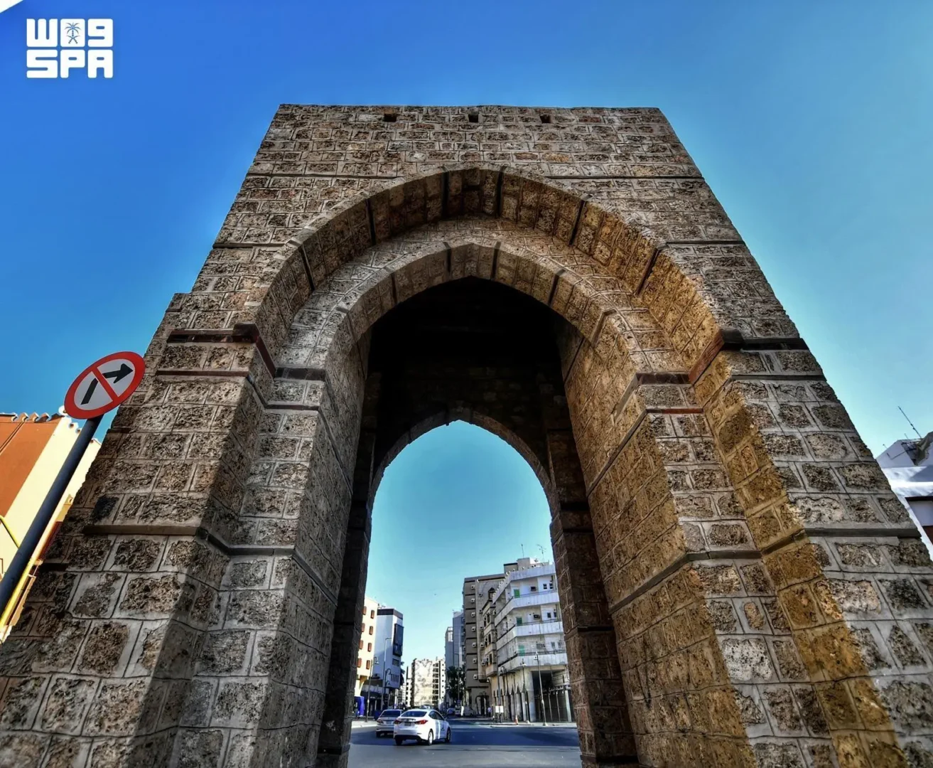Current image: Historic stone gateway with a large arched opening in Jeddah, with modern buildings and a road visible beyond it.