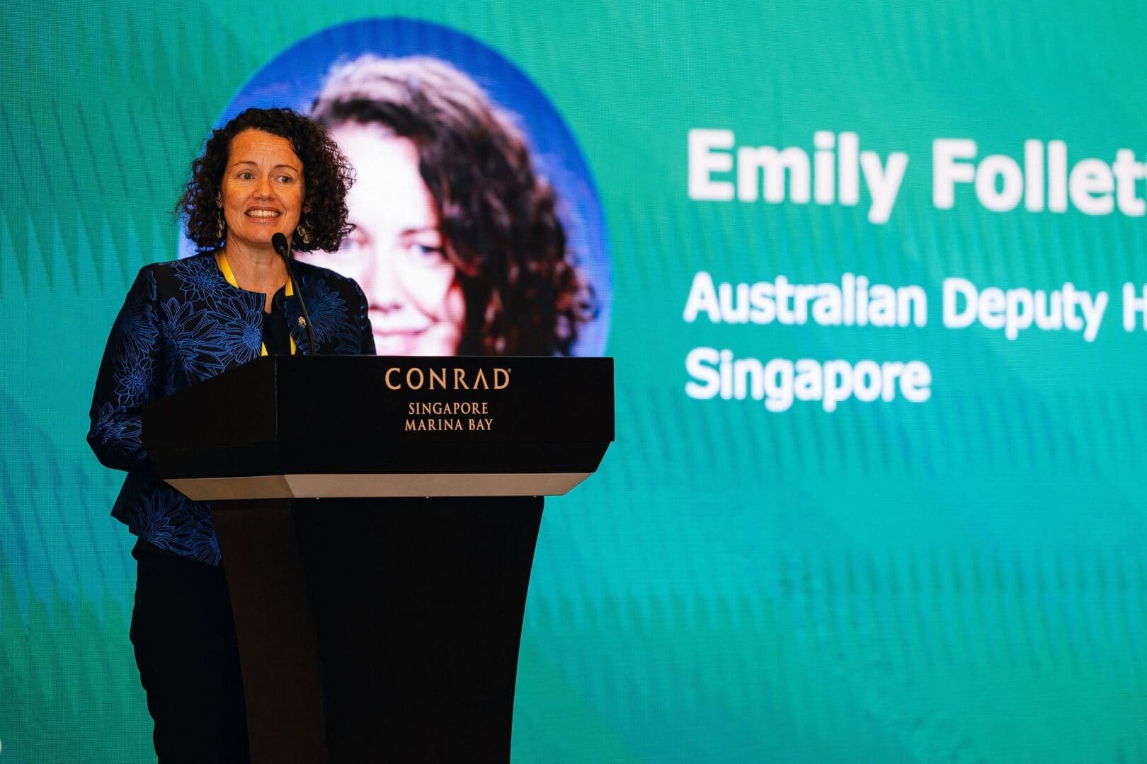Australian Deputy High Commissioner Emily Follett speaks from a podium during the opening address of an AI forum. The screen behind her identifies the session as “Australia and Singapore shaping the future of AI” and displays her name and title.